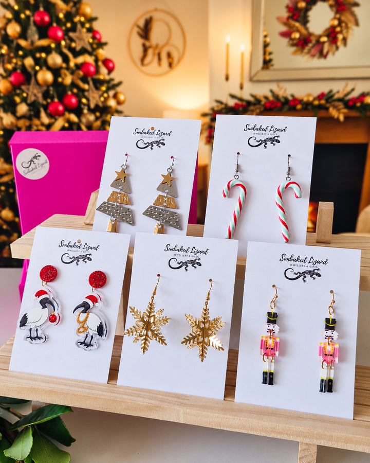 Statement Christmas Earrings Gift Box, five pairs of Christmas drop earrings on Sunbaked Lizard cards, including a gold snowflake and a nutcracker, in front of a decorated tree and fireplace