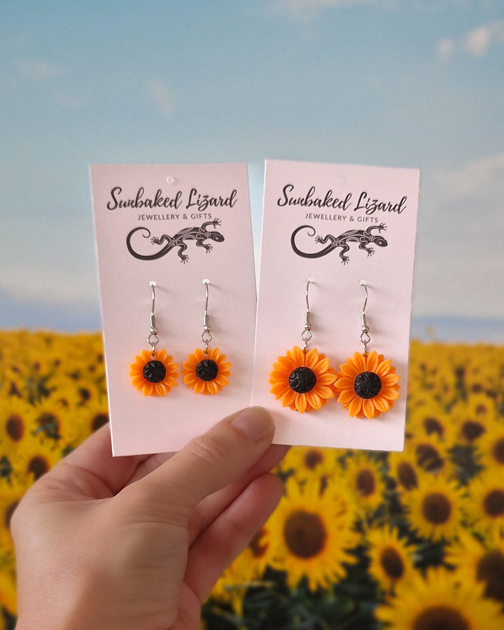 Photo showing the two different sizes of sunflower dangles earrings on display cards held against a sunflower field background.