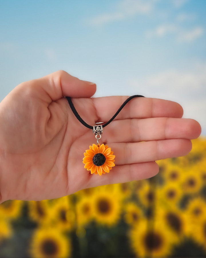 Hand holding a sunflower pendant necklace with a blurred sunflower field in the background