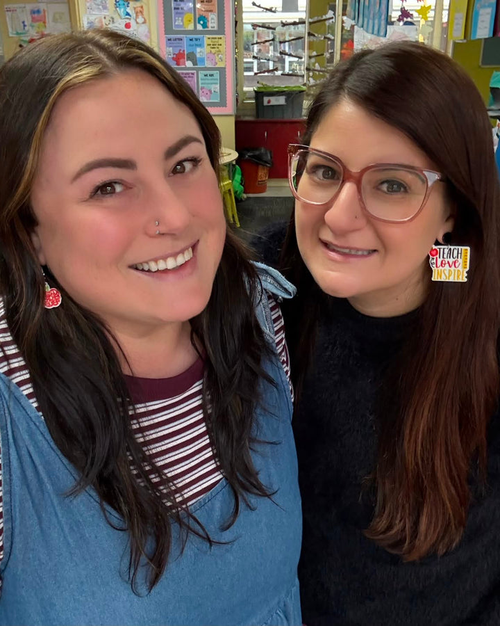 Two teachers from @two.bright.teachers wearing Sunbaked Lizard teacher earrings, including Teach Love Inspire and Best Teacher Ever designs, in a colourful classroom.