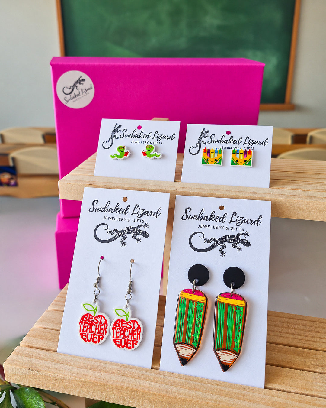 Teacher Thank You Earrings Gift Box, four pairs of classroom earrings on Sunbaked Lizard cards - a bookworm, crayons, a Best Teacher Ever apple, and a pencil - in front of a pink gift box and a chalkboard