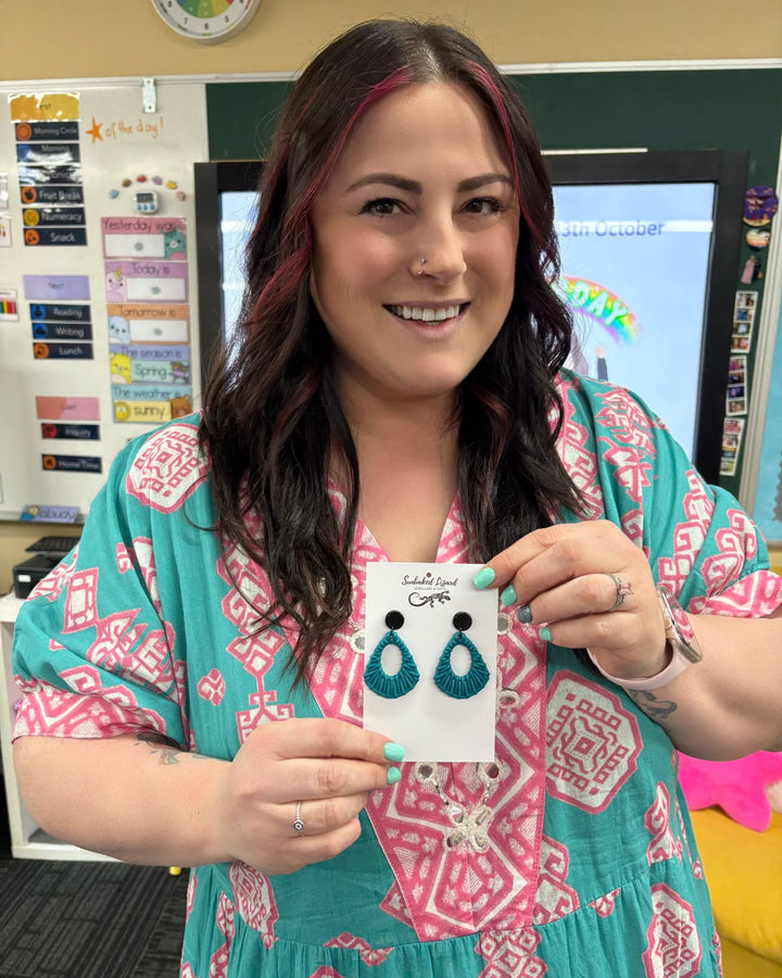 Teal Acrylic Woven Earrings with black tops displayed on a Sunbaked Lizard product card in a classroom.