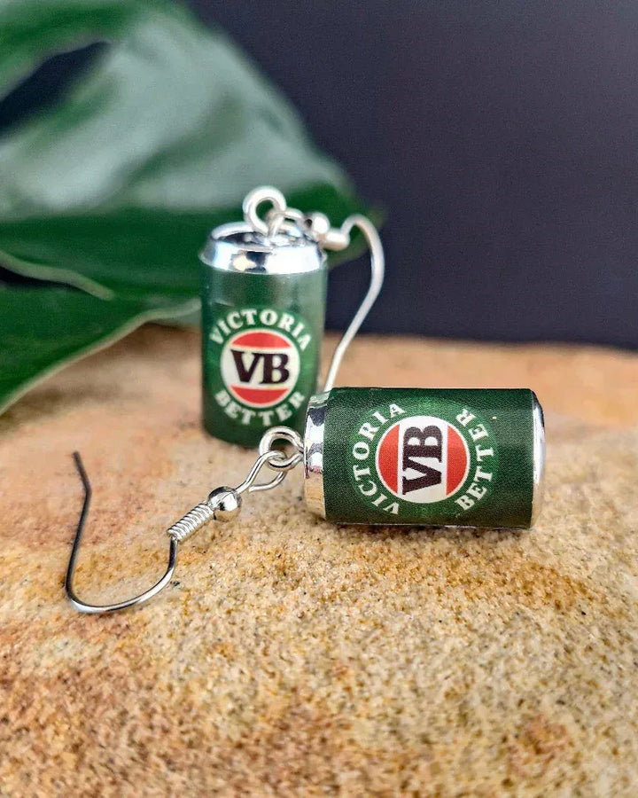 Close-up of Victoria Bitter VB beer can novelty earrings with silver hooks on sand background