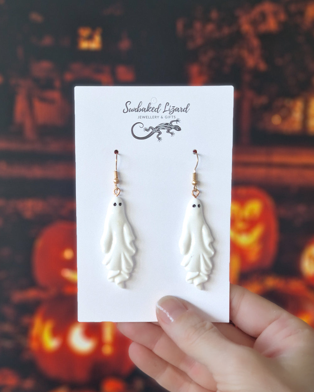 White ghost Halloween earrings displayed on a Sunbaked Lizard card showing scale with a spooky pumpkin Halloween background