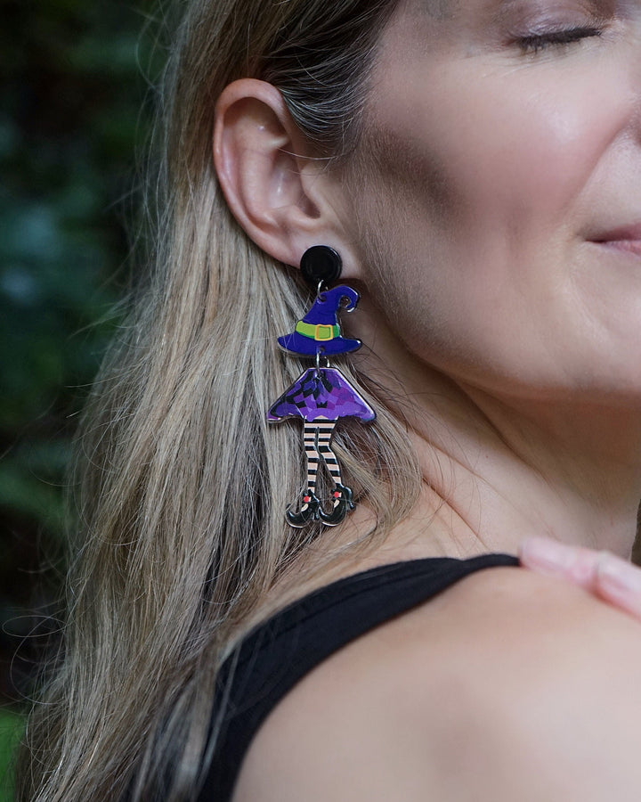 Close-up of a model wearing Witch Stud Drop Earrings with striped witch legs, a purple dress and pointed witch hat.