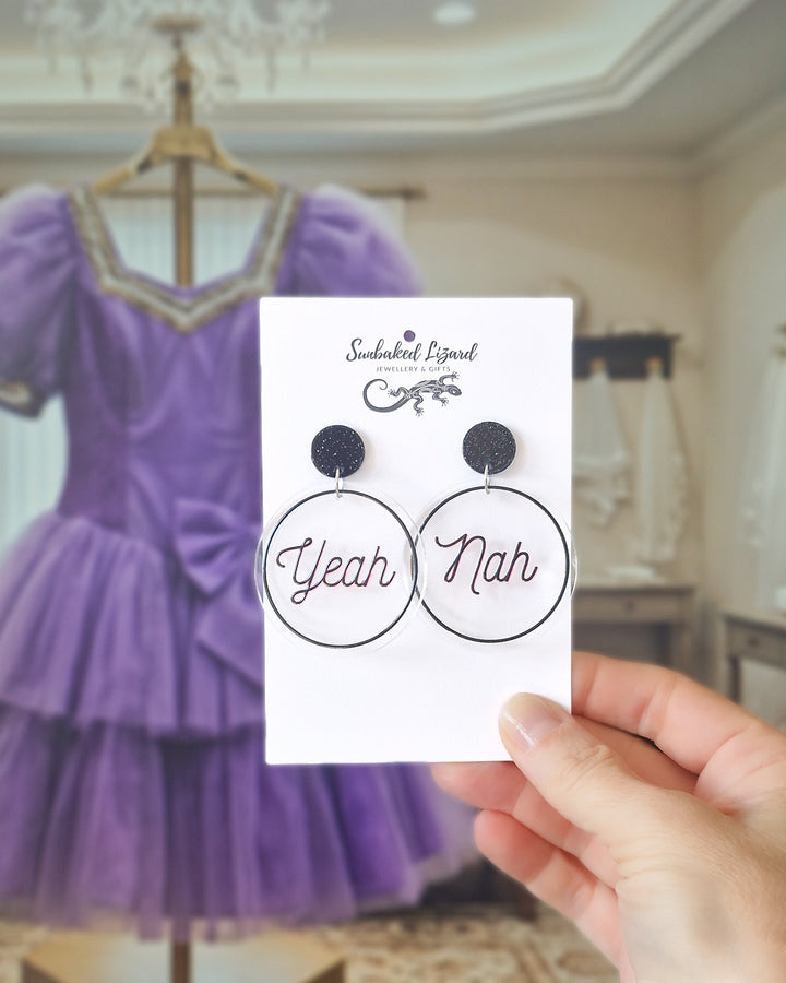 Yeah Nah black acrylic statement earrings displayed on a Sunbaked Lizard card with a purple dress behind.