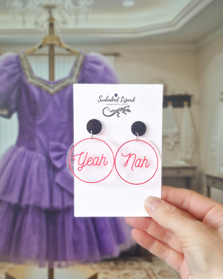 Yeah Nah pink acrylic statement earrings displayed on a Sunbaked Lizard card with a purple dress behind.