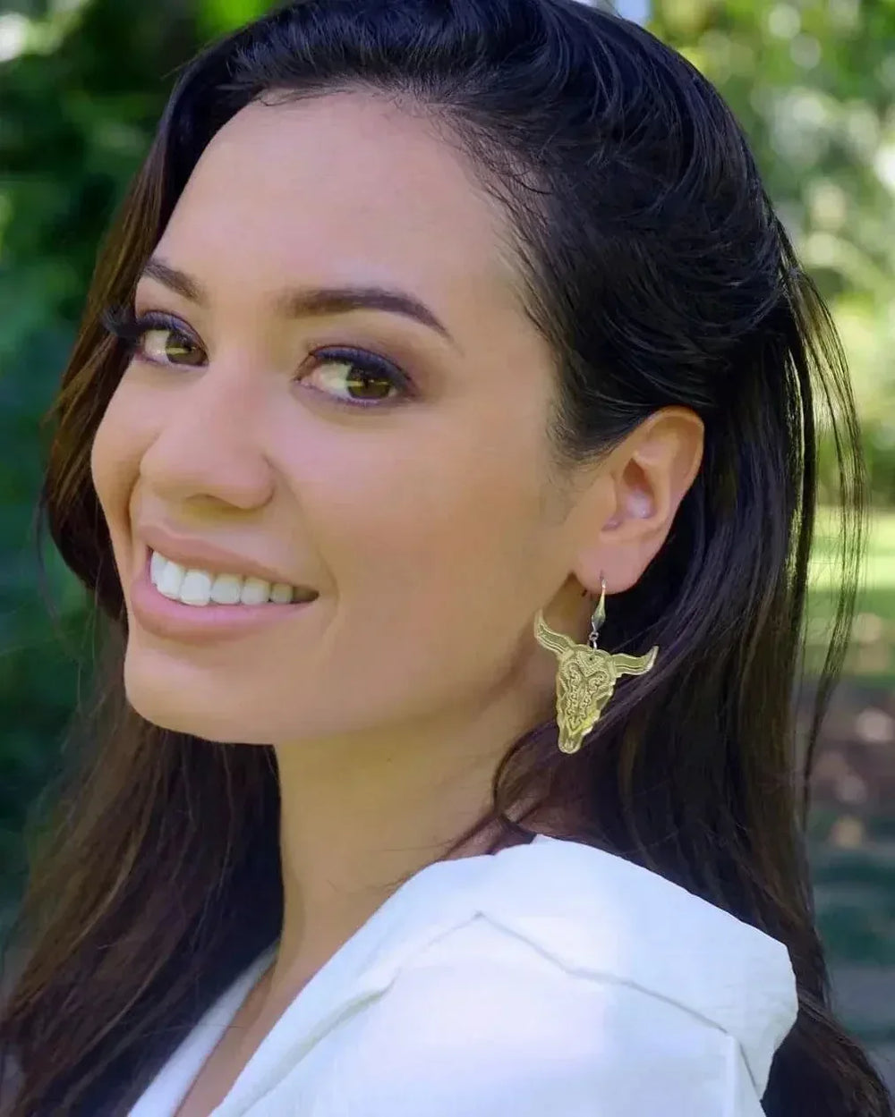 Close-up of woman wearing gold bull skull statement earrings outdoors in natural light.